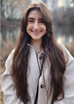 Headshot of Anna-Maria smiling at camera wearing cream coloured coat with long wavy brown hair falling in front of shoulders and face, standing in front of blurred nature background