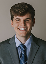 Headshot of William Frye smiling at camera in front of grey backdrop wearing blue checked tie and dark grey suit