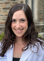 Headshot of student smiling at camera with dark brown curled hair wearing white coat in front of brick wall