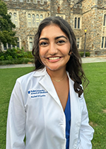 Student smiling at camera with brown curly hair wearing white coat over blue dress, standing on lawn in front of brick building