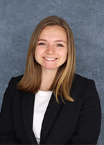 Headshot of student smiling at camera with blonde shoulder length hair wearing black blazer over white shirt in front of blue backdrop