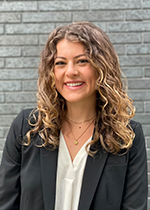 Student smiling at camera with light brown curly hair with blonde highlights, wearing black blazer over white shirt in front of grey brick wall