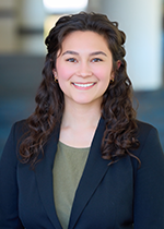 Headshot of student smiling at camera, with dark curls falling over both shoulders, wearing navy blazer over green top
