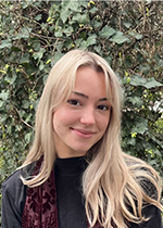 Student closed-mouth smiling at camera with light blonde hair falling over shoulder, wearing dark coloured shirt in front of greenery