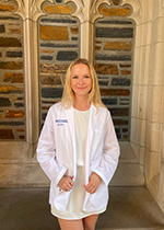 Student with blonde shoulder length hair holding edges of White Coat worn over cream dress in front of brick wall