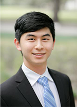 Student with black hair smiling at camera in front of blurred background wearing black suit, white shirt and blue striped tie