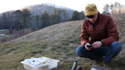 Julian Liber, a fourth-year PhD student in hat and puffy jacket, sitting on the side of a mountain collecting fungal samples.