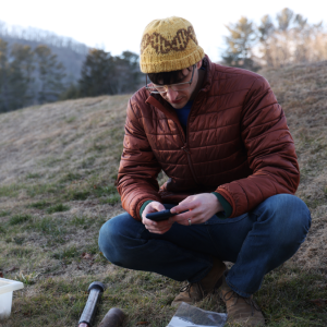 Male researcher in hat and puffy jacket, sitting on the side of a mountain collecting fungal samples. 